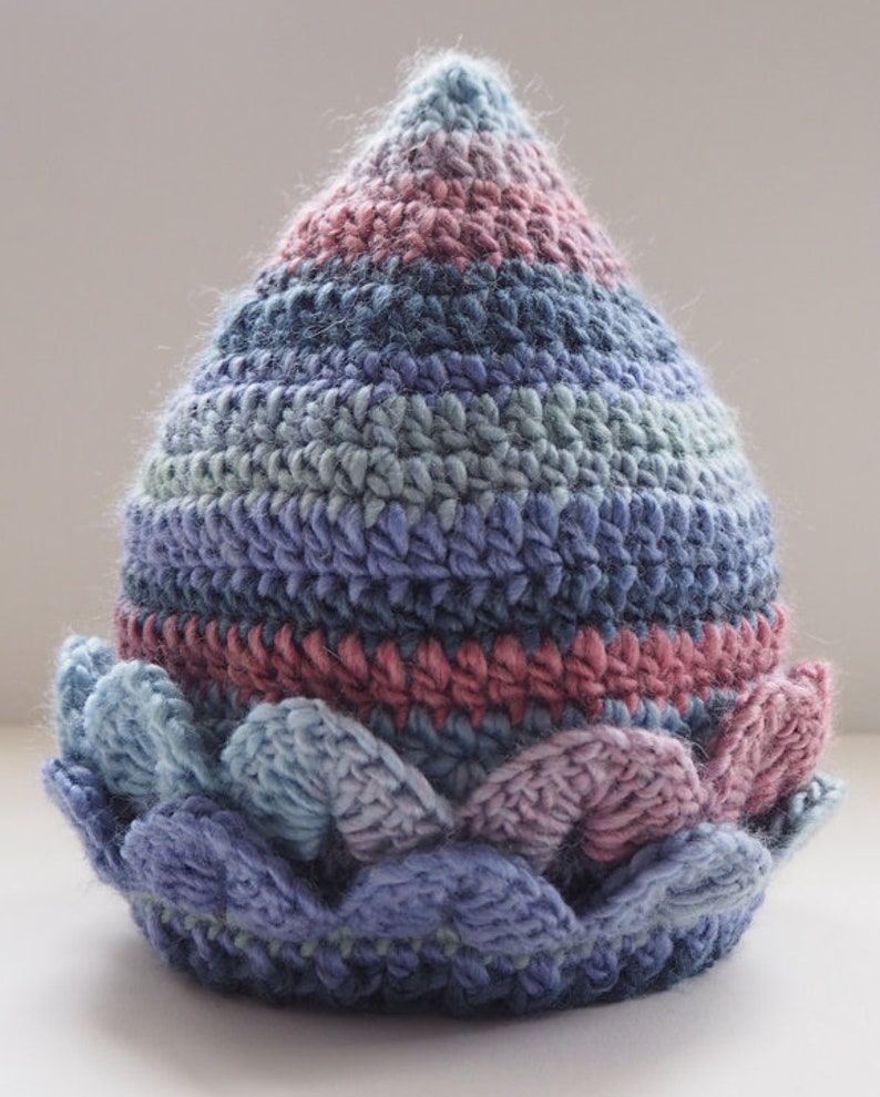 K&ouml;nnte beinhalten: Eine geh&auml;kelte M&uuml;tze mit einer spitzen Spitze und einem gewellten Rand. Die M&uuml;tze ist in Blaut&ouml;nen, Rosa und Gr&uuml;n gehalten.