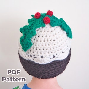 May include: A white and brown crocheted hat shaped like a Christmas pudding with green holly and red berries on top. The hat is being worn by a child. The text "PDF Pattern" is visible at the bottom of the image.
