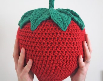 Puede incluir: Una almohada con forma de fresa hecha a crochet con una hoja verde de crochet en la parte superior. La almohada es roja y la hoja es verde.