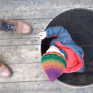 K&ouml;nnte beinhalten: Ein Kind mit einer bunten H&auml;kelm&uuml;tze und einer blauen Jacke schaut in die Kamera. Das Kind steht auf einer Holzoberfl&auml;che mit einem runden Loch in der Mitte.