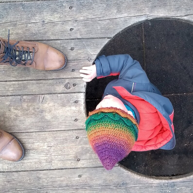 K&ouml;nnte beinhalten: Ein Kind mit einer bunten H&auml;kelm&uuml;tze und einer blauen Jacke schaut in die Kamera. Das Kind steht auf einer Holzoberfl&auml;che mit einem runden Loch in der Mitte.