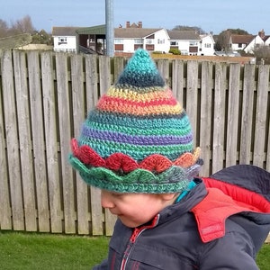 K&ouml;nnte beinhalten: Ein Kind tr&auml;gt eine bunte H&auml;kelm&uuml;tze mit einem gewellten Rand. Die M&uuml;tze ist in den Farben Gr&uuml;n, Blau, Lila, Rot und Gelb gestreift.