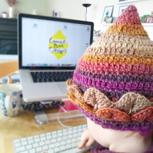 K&ouml;nnte beinhalten: Nahaufnahme einer Person, die einen mehrfarbigen H&auml;kelhut mit braunem Rand tr&auml;gt. Der Hut hat eine spitze Spitze und ist aus verschiedenen Farben gefertigt, darunter Pink, Lila, Orange und Braun. Die Person sitzt an einem Schreibtisch mit einem Laptop vor sich. Auf dem Laptop-Bildschirm ist eine gelbe Zitrone mit dem Text "Lemon Bee Designs" zu sehen.