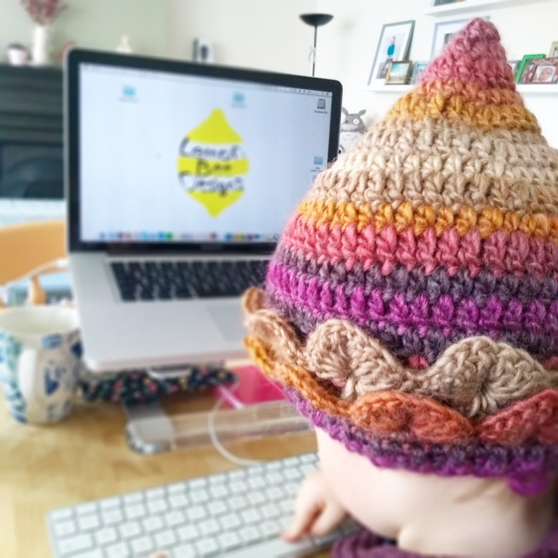 K&ouml;nnte beinhalten: Nahaufnahme einer Person, die einen mehrfarbigen H&auml;kelhut mit braunem Rand tr&auml;gt. Der Hut hat eine spitze Spitze und ist aus verschiedenen Farben gefertigt, darunter Pink, Lila, Orange und Braun. Die Person sitzt an einem Schreibtisch mit einem Laptop vor sich. Auf dem Laptop-Bildschirm ist eine gelbe Zitrone mit dem Text "Lemon Bee Designs" zu sehen.