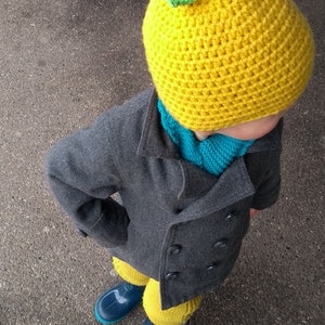 Puede incluir: Un ni&ntilde;o con un gorro de lim&oacute;n amarillo tejido a crochet con una hoja verde en la parte superior, un abrigo gris, una bufanda azul y botas de lluvia azules.