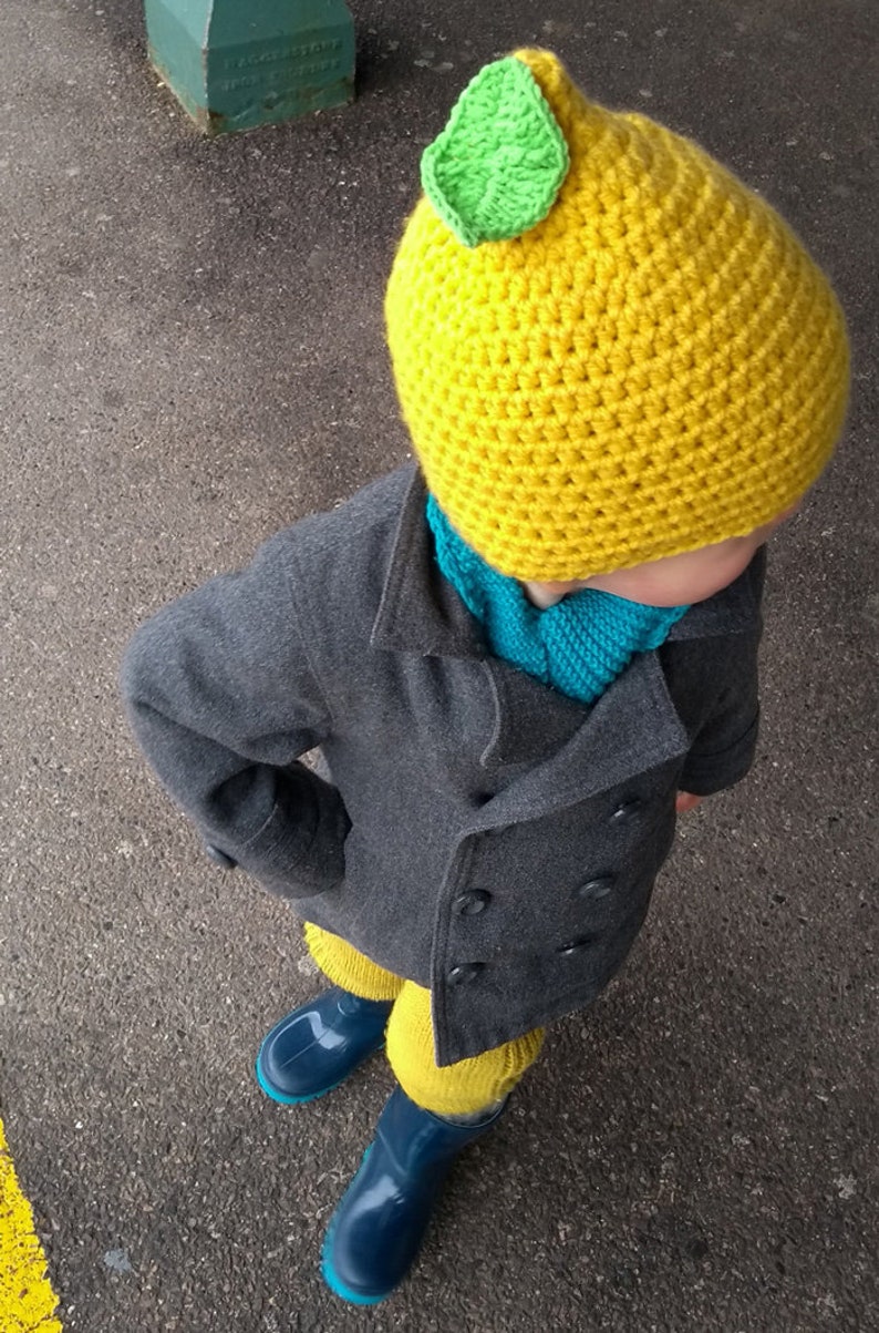 Puede incluir: Un ni&ntilde;o con un gorro de lim&oacute;n amarillo tejido a crochet con una hoja verde en la parte superior, un abrigo gris, una bufanda azul y botas de lluvia azules.