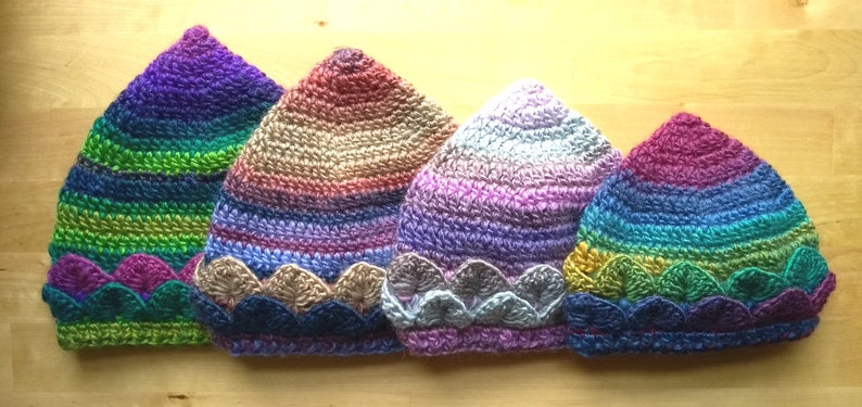 K&ouml;nnte beinhalten: Vier bunte H&auml;kelm&uuml;tzen mit einem gewellten Rand. Die M&uuml;tzen sind in den Farben Lila, Gr&uuml;n, Rosa und Blau.