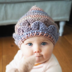 K&ouml;nnte beinhalten: Ein Baby tr&auml;gt eine geh&auml;kelte M&uuml;tze mit einem gewellten Rand in den Farben Lila, Rosa und Braun.