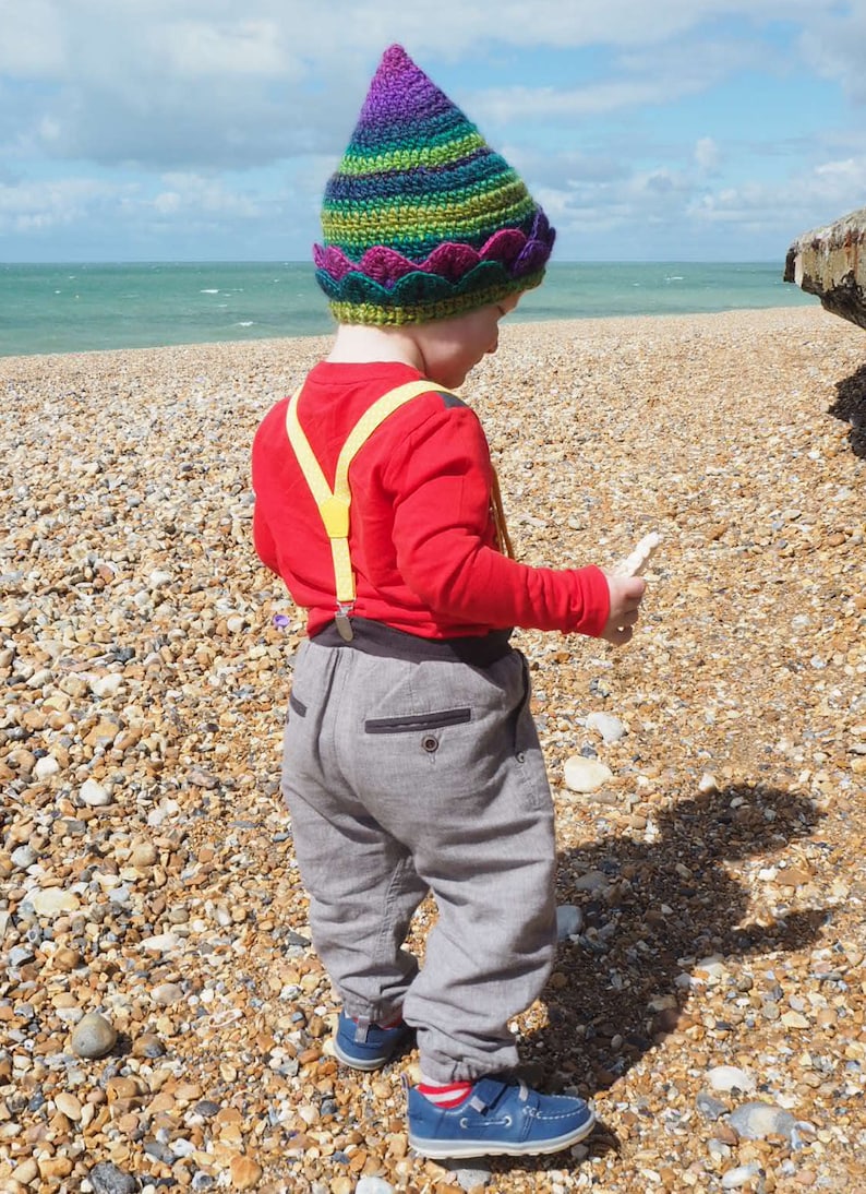 K&ouml;nnte beinhalten: Ein kleines Kind tr&auml;gt eine bunte H&auml;kelm&uuml;tze mit einer spitzen Spitze und einem gewellten Rand. Das Kind tr&auml;gt ein rotes Langarmshirt, graue Hosen und gelbe Hosentr&auml;ger. Das Kind steht an einem Strand mit kleinen Kieselsteinen.