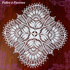 Pode incluir: Uma toalha de mesa quadrada branca de crochê com um intrincado design floral. A toalha de mesa apresenta um design circular central rodeado por intrincados motivos em forma de folha e uma borda ondulada. O texto "Folies à Facettes" está no canto superior esquerdo.