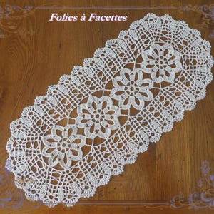 Peut inclure: Un napperon en crochet blanc de forme ovale présentant un motif floral répétitif avec des points ajourés complexes et un bord festonné. Le texte "Folies à Facettes" est visible en haut au centre de l'image au-dessus de la pièce textile.