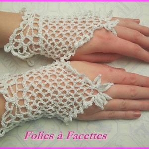 Peut inclure: Une paire de mitaines en crochet blanc à mailles ajourées. Les gants présentent un motif floral délicat sur les bords. Le texte "Folies à Facettes" est visible en bas. Les gants sont présentés sur un fond clair.