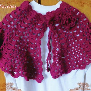 Peut inclure: Une capelette en crochet magenta présentant un motif ajouré en forme de coquilles et une fleur en crochet décorative sur le côté droit. Le vêtement est ouvert sur le devant et est présenté sur un haut blanc. Le texte Folies à Facettes est visible.