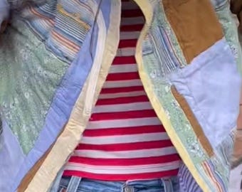 May include: A blue and yellow patchwork jacket with a zipper closure. The jacket is worn over a red and white striped shirt and blue jeans.