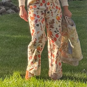 May include: Cream-colored pants with a vibrant fruit and floral pattern. The pants feature a wide-leg design and are paired with a matching jacket. The person is wearing a cream-colored sweater and brown belt. The outfit is set against a backdrop of green grass.