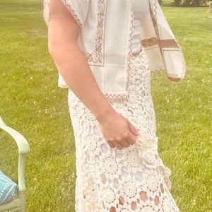 May include: Cream-colored crochet skirt and matching short-sleeved jacket with floral embroidery. The outfit is worn over a white tank top. The skirt features a scalloped hem and a floral pattern. The jacket has a collar and decorative trim.