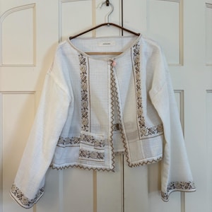 May include: A white jacket with long sleeves, featuring intricate floral embroidery in shades of brown and beige. The jacket has a scalloped hem and a small pocket. The jacket is hanging on a wooden hanger.