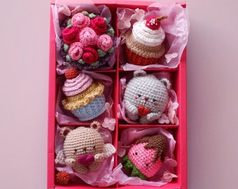 May include: A red box filled with six crocheted amigurumi toys. The toys are shaped like cupcakes, a strawberry, and bears. The box is lined with pink tissue paper.