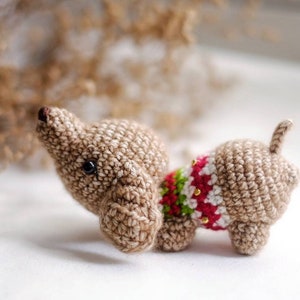 Puede incluir: Un pequeño dachshund de crochet que lleva un suéter rojo, verde y blanco con cuentas doradas. El dachshund es marrón y tiene ojos negros.