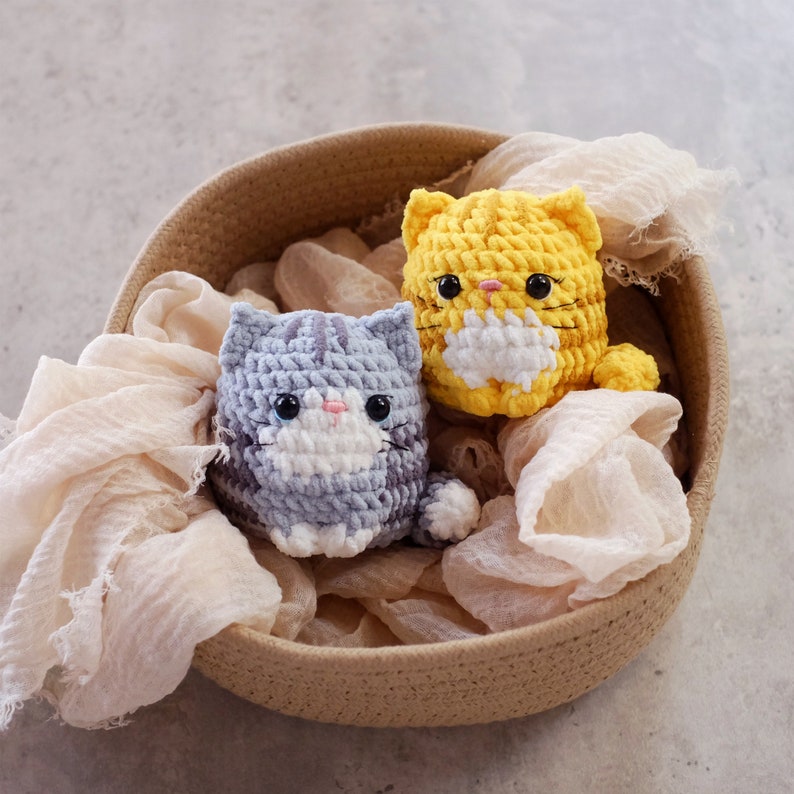Puede incluir: Dos juguetes de crochet con forma de gato en una cesta tejida. Un gato es amarillo y el otro es gris. Ambos gatos tienen la cara blanca y los ojos negros.