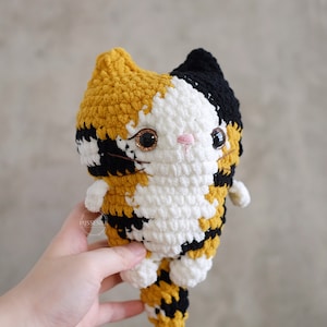 May include: A crocheted calico cat plush toy with black, white, and yellow yarn. The cat has large, sparkly eyes and a pink nose.