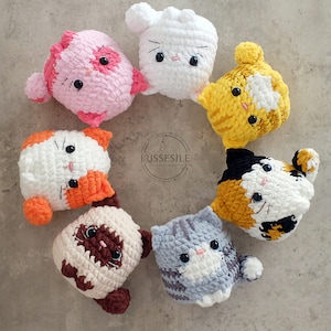 Puede incluir: Una colecci&oacute;n de ocho peluches de gato de crochet en varios colores, incluyendo rosa, blanco, amarillo, naranja, marr&oacute;n y gris. Cada peluche tiene una expresi&oacute;n facial &uacute;nica y presenta ojos y nariz bordados.