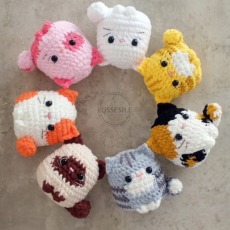 Puede incluir: Una colecci&oacute;n de ocho peluches de gato de crochet en varios colores, incluyendo rosa, blanco, amarillo, naranja, marr&oacute;n y gris. Cada peluche tiene una expresi&oacute;n facial &uacute;nica y presenta ojos y nariz bordados.