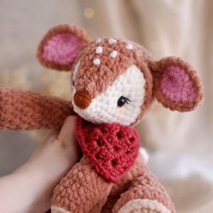 May include: A handmade crochet deer plushie with a heart-shaped bib. The deer features a brown body, white spots, and pink-lined ears. The plushie is soft and cuddly, perfect for a child's toy or nursery decor.