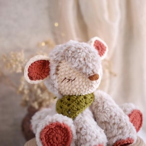 May include: A handmade crochet teddy bear with a cream-colored body and rust-colored accents on the ears and feet. The bear has a green scarf and closed eyes, giving it a peaceful appearance.