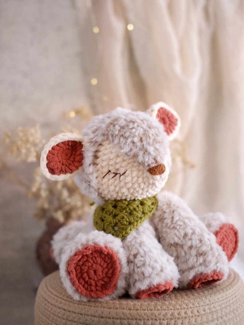 May include: A handmade crochet teddy bear with a cream-colored body and rust-colored accents on the ears and feet. The bear has a green scarf and closed eyes, giving it a peaceful appearance.