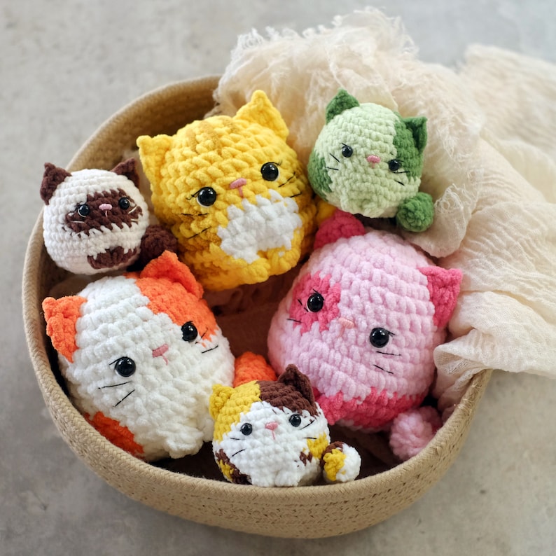 Puede incluir: Una colecci&oacute;n de seis juguetes de crochet con forma de gato en una cesta de mimbre. Los gatos son de varios colores, incluyendo rosa, amarillo, verde y blanco.