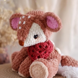May include: A handmade crochet deer plushie in shades of brown, white, and red. The deer has white spots, pink-lined ears, and a red scarf. The plushie is sitting on a woven surface.