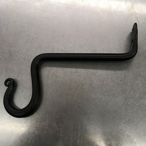 May include: Black metal hook with a curved design. The hook is attached to a wall or surface.