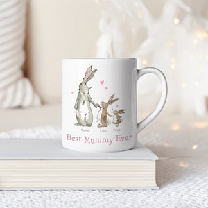 Pu&ograve; includere: Una tazza in ceramica bianca con un design di tre conigli che si tengono per mano. I conigli sono etichettati "Mummy", "Elsie" e "Pippa". Il testo "Best Mummy Ever" &egrave; stampato in rosa sotto i conigli.