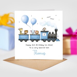 May include: A birthday card featuring a blue train with animal passengers and two blue balloons. The train has a monkey, giraffe, tiger, elephant, and lion. Text reads "Happy 1st Birthday to choo! To a very special Son Thomas."