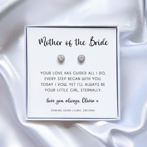 May include: A white gift box featuring the text "Mother of the Bride" and two sparkling stud earrings. The box contains a message and the words "Sterling Silver | Cubic Zirconia". The background is a white, silky fabric.