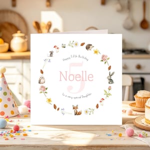 May include: A square white birthday card featuring a large pink number five and the name Noelle in the center. A circular wreath of watercolor illustrations surrounds the text, including a fox, rabbit, owl, hedgehog, mouse, flowers, and pinecones.