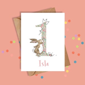 May include: A birthday card with a floral number one and a cartoon rabbit wearing a crown. The rabbit is dancing next to the number one. The name "Isla" is written in pink script. The card is inside a brown envelope.