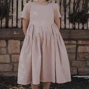 Może przedstawiać: Jasnoróżowa sukienka z krótkimi rękawami i marszczoną spódnicą. Sukienka ma okrągły dekolt i sięga do połowy łydki. Osoba ma na sobie buty we wzory. Tło obejmuje białe ogrodzenie i ceglany mur.