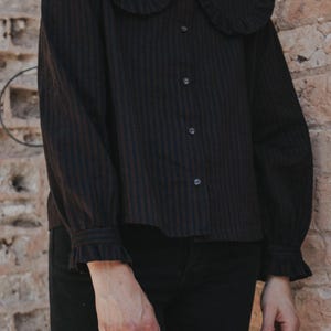Puede incluir: Una camisa negra de manga larga con finas rayas verticales oscuras, que presenta un cuello grande y redondeado con bordes con volantes, cierre frontal de botones y pu&ntilde;os a juego con volantes en las mu&ntilde;ecas, combinada con pantalones negros.