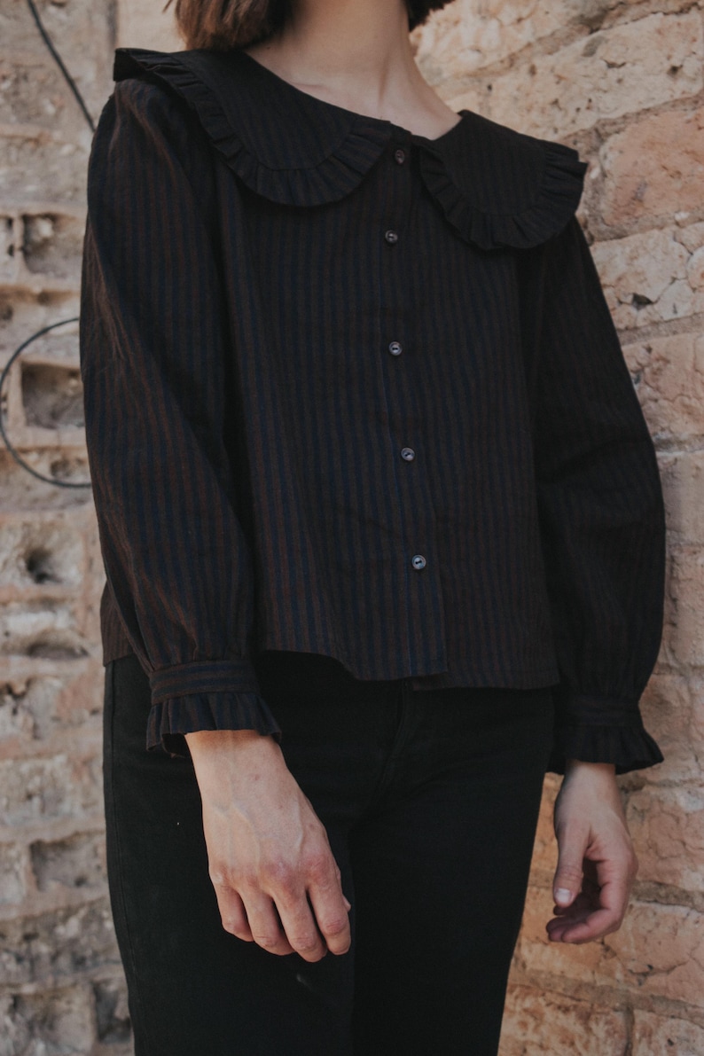 Puede incluir: Una camisa negra de manga larga con finas rayas verticales oscuras, que presenta un cuello grande y redondeado con bordes con volantes, cierre frontal de botones y pu&ntilde;os a juego con volantes en las mu&ntilde;ecas, combinada con pantalones negros.
