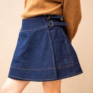 May include: A dark blue denim wrap skirt featuring a high-waisted design with two adjustable side straps and metallic buckles. The skirt has visible orange contrast stitching along the hem and seams, creating a structured A-line silhouette.