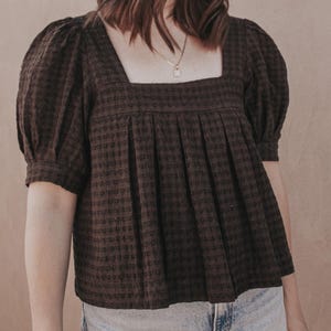 Peut inclure: Un chemisier marron foncé avec une encolure carrée, des manches courtes bouffantes et un motif à carreaux texturé. Le vêtement présente un corsage avant plissé créant une silhouette ample et fluide, associé à un jean bleu clair.