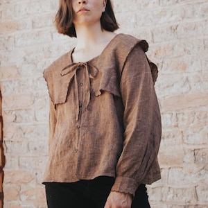 Könnte beinhalten: Eine braune Langarmbluse mit Rüschenkragen und Bindverschluss. Die Bluse hat ein vertikales Streifenmuster und Knöpfe. Die Ärmel haben gerüschte Manschetten. Die Bluse wird mit einer schwarzen Hose kombiniert. Der Hintergrund ist eine Backsteinmauer.