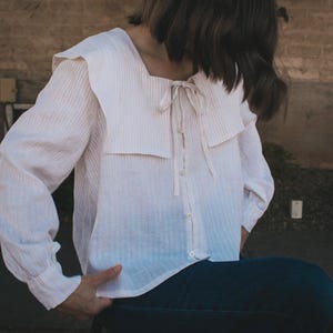 Może przedstawiać: Jasna, pasiasta bluzka z dużym, kwadratowym kołnierzem i długimi rękawami. Bluzka ma zapinany na guziki przód i wiązanie przy dekolcie. Tkanina wydaje się być lnem lub podobnym lekkim materiałem.