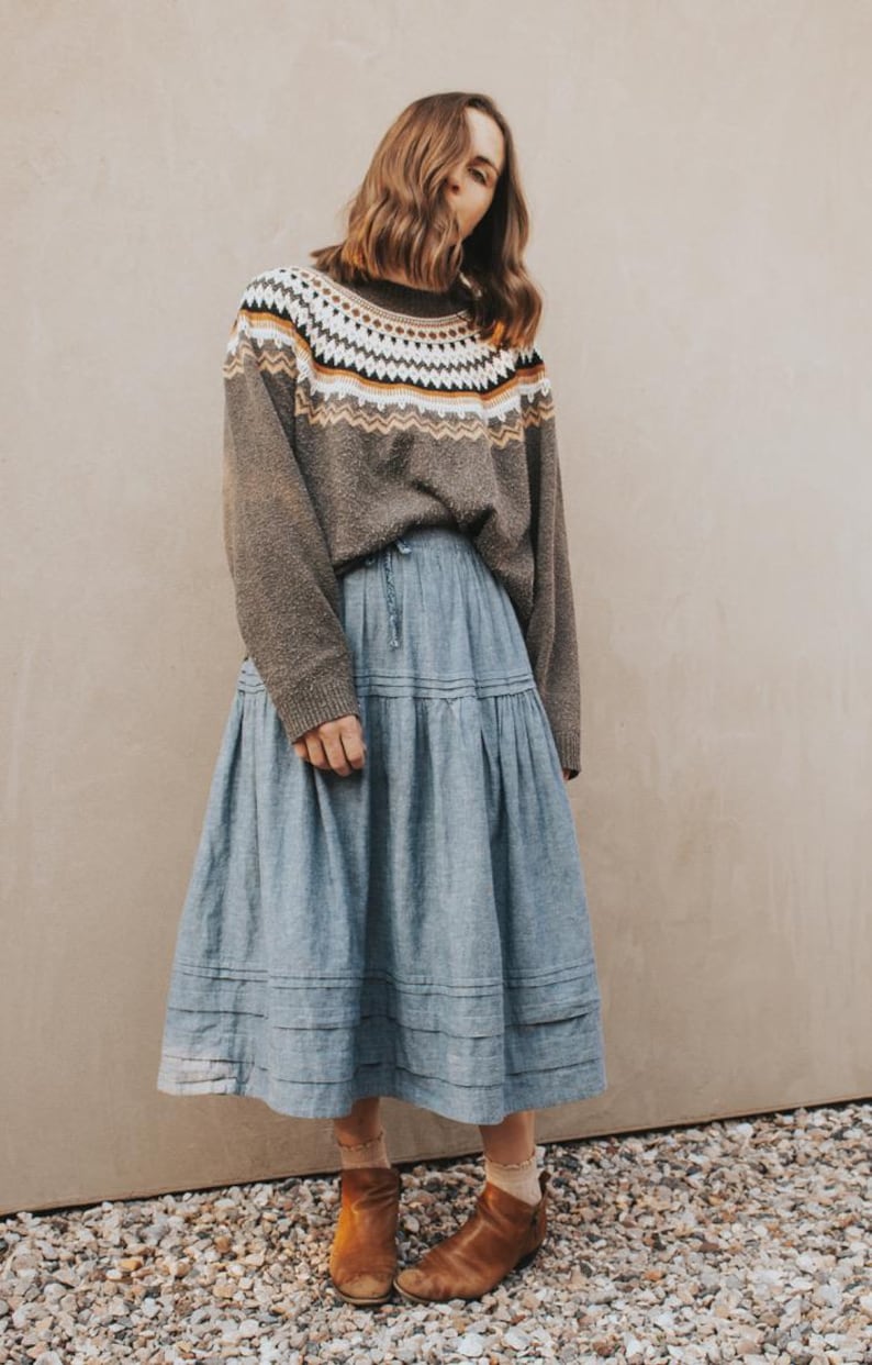 May include: A woman wearing a brown sweater with a geometric pattern in white, yellow, and orange. She is also wearing a light blue tiered skirt and brown ankle boots. The outfit is casual and stylish.