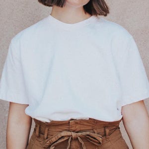 Puede incluir: Una camiseta blanca de gran tamaño con un cinturón marrón de cintura alta. El cinturón tiene un detalle de lazo.