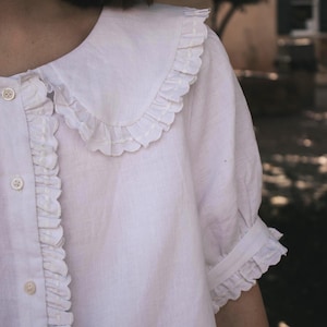 Puede incluir: Una blusa blanca de manga corta con un cuello grande y redondeado con bordes con volantes, una tapeta frontal con botones con ribete de volantes a juego y mangas terminadas con un detalle de pu&ntilde;o con volantes fruncidos.