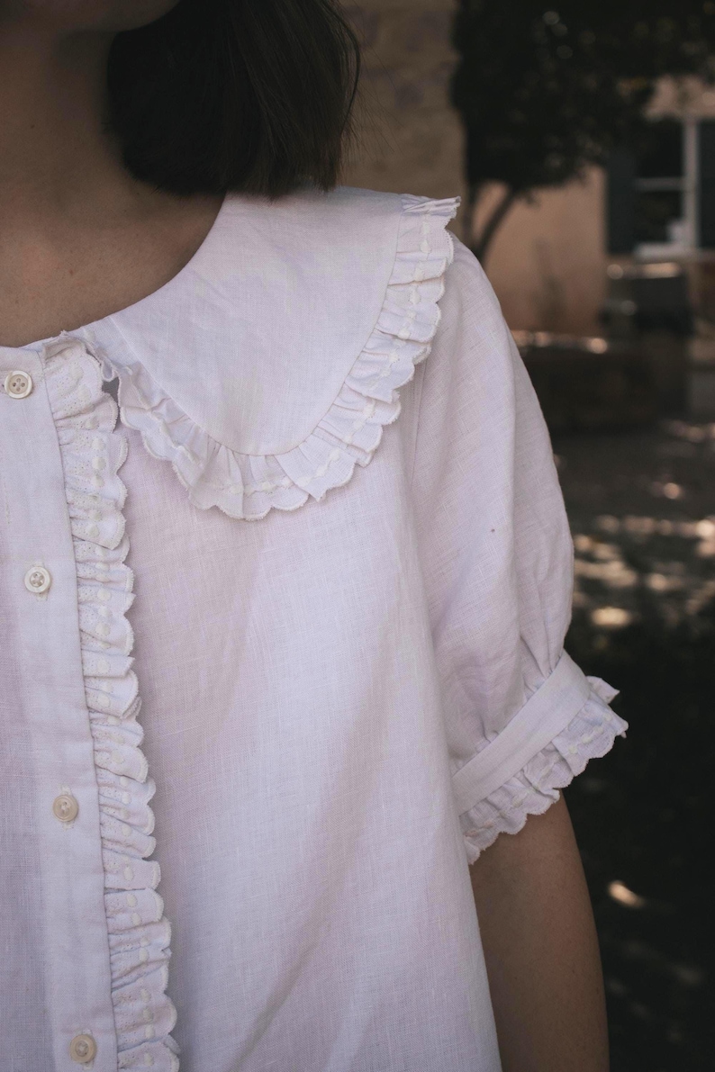Puede incluir: Una blusa blanca de manga corta con un cuello grande y redondeado con bordes con volantes, una tapeta frontal con botones con ribete de volantes a juego y mangas terminadas con un detalle de pu&ntilde;o con volantes fruncidos.