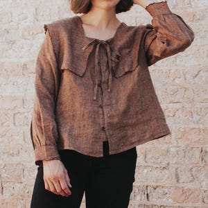 Könnte beinhalten: Ein braunes, gestreiftes Leinenhemd mit einem großen, gekräuselten Kragen und einer Schleife am Hals. Das Hemd hat einen Knopfleisten vorne und lange Ärmel.